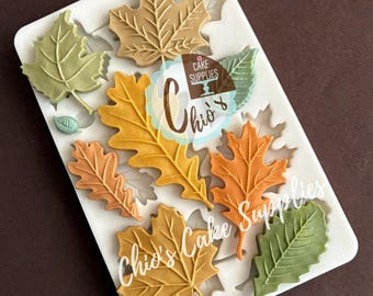 May include: A white silicone mold featuring various autumn leaf designs in shades of green, orange, and brown. The mold includes maple and oak leaf shapes, perfect for creating decorative elements for cakes and other baked goods. The text "Chio's Cake Supplies" is visible.