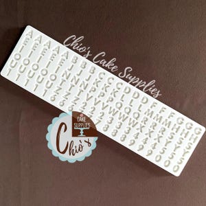 Könnte beinhalten: Weiße Plastikform mit erhabenen Buchstaben und Zahlen zum Backen und Dekorieren von Kuchen. Die Form enthält das Alphabet, die Zahlen 0-9 und das Und-Zeichen. Die Form ist mit "Chio's Cake Supplies" beschriftet.