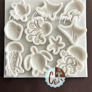 May include: A white silicone mold featuring various sea creatures, including a turtle, starfish, dolphin, octopus, jellyfish, and seashell. The mold has the text "Chio's Cake Supplies" in a script font. A circular logo with the text "Chio's Cake Supplies" is also visible.
