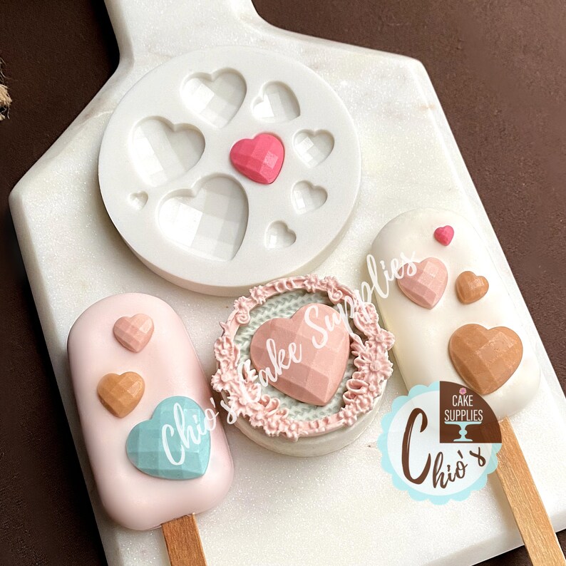 May include: A silicone mold with multiple heart shapes, a pink heart-shaped cake pop with a decorative heart on top, and a white cake pop with a brown heart on top.  The cake pops are on wooden sticks.  The cake pops are decorated with a variety of colors and designs.  The image also includes a pink and white cake pop with a decorative heart on top.  The cake pops are all on a white surface.  The image also includes the text "Chio's Cake Supplies".