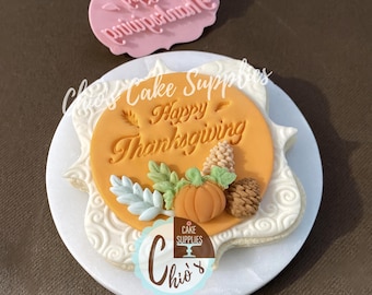 May include: A decorated cookie with an orange circle that says "Happy Thanksgiving" with a pumpkin, leaves, and pinecones. The cookie is on a white plate with a floral design. A pink stamp with text is above the cookie.
