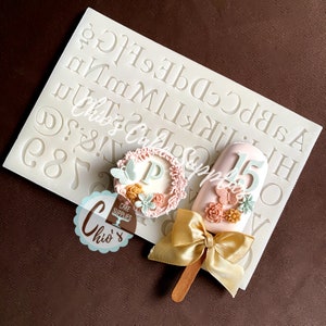 May include: A silicone mould with letters and numbers, a pink and white cake pop with a butterfly and flowers, and a pink and white cake pop with a butterfly and flowers and the number 15.