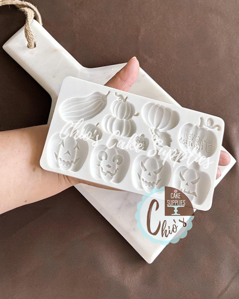 May include: A white silicone mold featuring various pumpkin designs and an eggplant. The mold is held over a white marble cutting board. The text "Chio's Cake Supplies" is printed on the mold. The mold is designed for cake decorating.