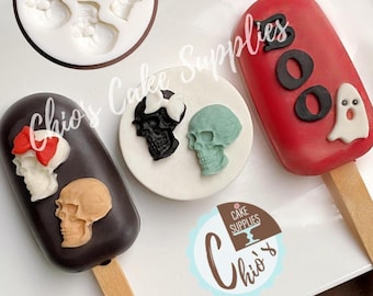May include: A collection of Halloween-themed cake pops and skull-shaped molds. The cake pops are decorated with skull designs, a ghost, and the word "BOO". The molds are white and the cake pops are various colors.