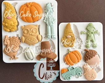 May include: Two white silicone molds filled with various fall-themed shapes. One mold features a gnome, pumpkin, scarecrow, owl, turkey, pilgrim hat, and a gourd. The other mold includes a gnome, scarecrow, pumpkin, acorn, and a truck carrying pumpkins.