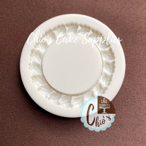 Puede incluir: Molde de silicona blanco para hornear un pastel con un borde decorativo de hojas. El molde es redondo y tiene una superficie lisa. El molde está diseñado para crear un pastel con un borde decorativo de hojas.