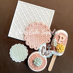 May include: A set of four silicone cake molds featuring a delicate lace pattern. The molds are in various sizes and shapes, including a square, a circle, and a popsicle mold. The molds are white, pink, and green.