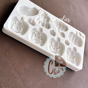 May include: A white silicone mold featuring various Halloween-themed shapes, including pumpkins and gourds. The mold has multiple cavities for creating decorative elements. The text "Chio's Cake Supplies" is visible on the mold.