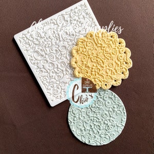 May include: Three silicone molds for baking and decorating cakes. The molds are shaped as a square, a circle, and a scalloped circle. The molds have a delicate, intricate design of swirls and vines.