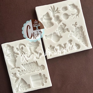 May include: Two white silicone molds for cake decorating featuring various holiday-themed shapes including a character, a candy cane, a gift box, a Christmas tree, and the text MERRY CHRISTMAS. The molds have multiple recessed cavities.