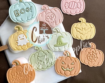 May include: A collection of Halloween-themed pumpkin-shaped decorations in various pastel colors. Some pumpkins feature designs like a skull, cat, witch hat, and the text "BOO!" and "HAPPY HALLOWEEN".