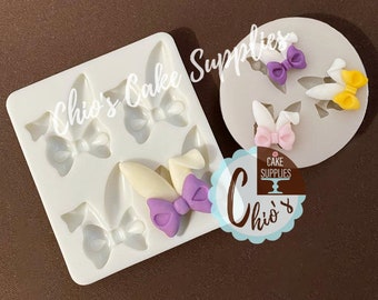 May include: A white silicone mould featuring bunny ear and bow shapes, with a circular mould containing finished bunny ear and bow decorations in purple, pink, and yellow. The image includes the text "Chio's Cake Supplies".