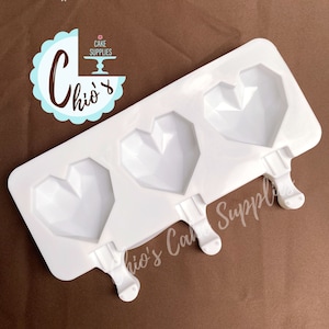 May include: White silicone mould with three heart-shaped cavities. Each cavity has a stick attached for making popsicles. The mould is on a brown surface.