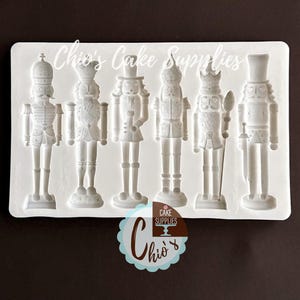 May include: A white silicone mold featuring six detailed nutcracker soldier designs. The mold includes the text "Chio's Cake Supplies". The nutcrackers are in various poses, with intricate uniform details, perfect for cake decorating.