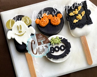 May include: A collection of Halloween-themed cake pops on a white marble surface. The pops feature designs like a Mickey Mouse ghost, pumpkin faces, bats, and a spider with the word "BOO". Each pop is on a wooden stick.