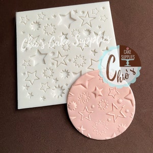 May include: A white silicone mold with a design of various sized stars. A pink fondant imprint shows the design of the mold.