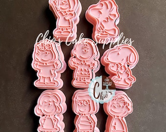 May include: Pink plastic cookie cutters featuring characters from the Peanuts comic strip. The set includes Snoopy, Charlie Brown, Lucy, and other characters. The text "Chio's Cake Supplies" is visible.