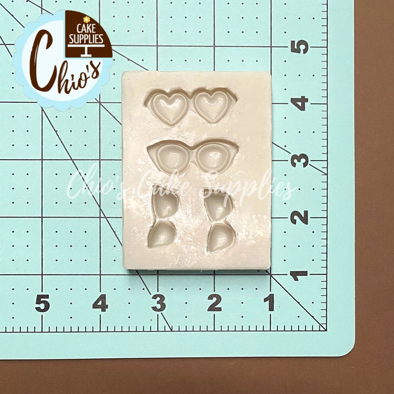 May include: A light-colored silicone mold featuring three sets of shapes: heart-shaped glasses, cat-eye glasses, and two pairs of leaf-shaped designs. The mold is on a blue and green grid background with the "Chio's Cake Supplies" logo.