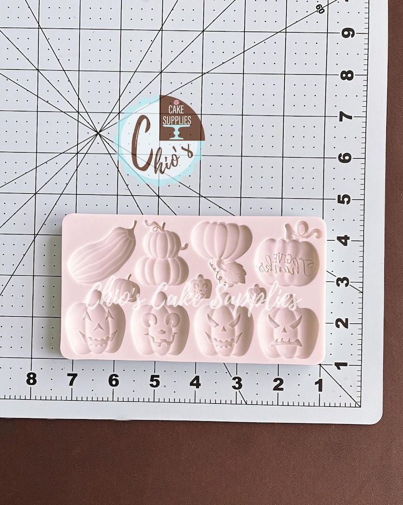 May include: A light pink silicone mold featuring various Halloween-themed shapes. The mold includes pumpkins, a gourd, and the text "GIVE THANKS". The mold is placed on a white and black grid with inch measurements.