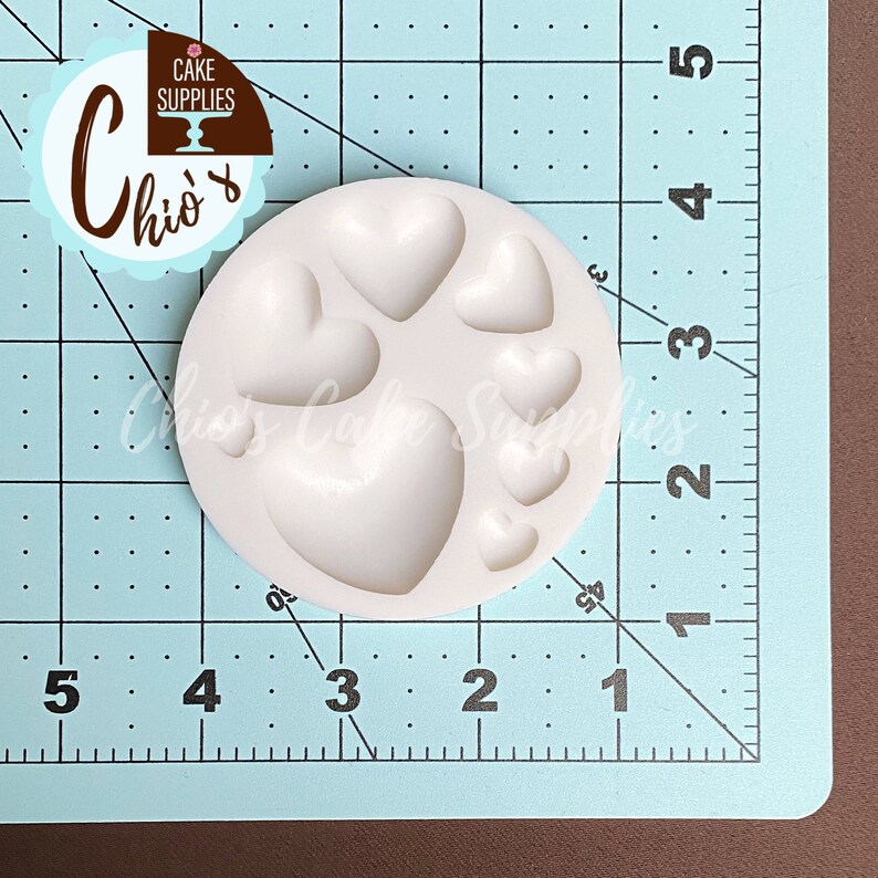 May include: White silicone mold with six heart-shaped cavities. The mold is on a blue gridded surface.