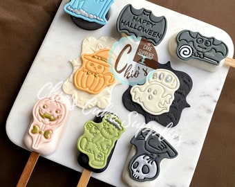 May include: A set of eight Halloween-themed cake pops. The pops are decorated with various Halloween characters, including a ghost, a pumpkin, a bat, a witch, a Frankenstein, a crow, and a tombstone. The pops are on wooden sticks and are arranged on a white marble surface.