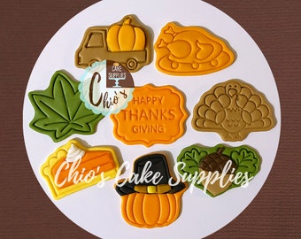 May include: A collection of Thanksgiving-themed cookie cutters in various shapes, including a pumpkin in a truck, a turkey, a leaf, a pie, and a pumpkin wearing a pilgrim hat. The words "Happy Thanksgiving" are also present. The cookie cutters are in shades of brown, orange, and green.
