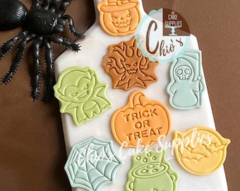 May include: A collection of Halloween-themed cookie cutters in various shapes, including a witch's pumpkin, a vampire, a grim reaper, and a spider web. The cutters are in shades of orange, green, and blue, arranged on a white cutting board.