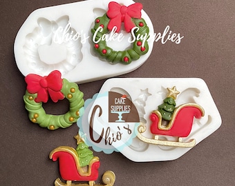 May include: Silicone mold for making Christmas-themed decorations. The mold features two wreaths with red bows and a sleigh with a Christmas tree. The mold is white and has a smooth surface.