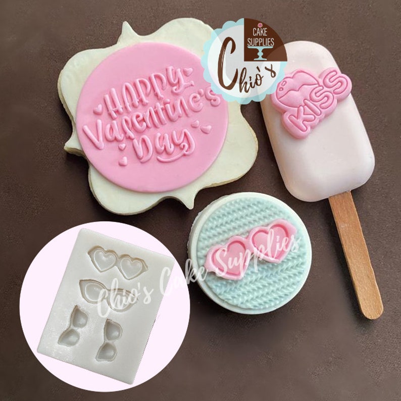 May include: A collection of Valentine's Day themed cake decorations. Includes a pink "Happy Valentine's Day" embossed circle, a pink popsicle with "KISS" text, a mold with heart-shaped glasses, and a cake with heart-shaped glasses.