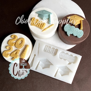 May include: A white silicone mold with two graduation caps, a pen, and the word "GRAD" in raised lettering. There are three decorated cookies in the background, one with the numbers "2024", one with the word "GRAD" and a graduation cap, and one with the words "Congratulations!" and a graduation cap.