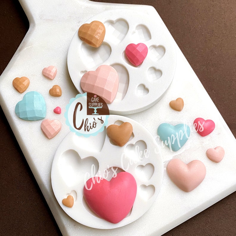 May include: A silicone mold for making heart-shaped fondant decorations. The mold has multiple heart-shaped cavities of different sizes. There are also several finished fondant hearts in various colors, including pink, blue, and brown.