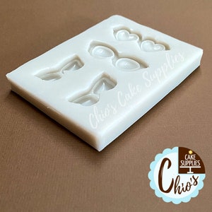 May include: A white silicone mold for cake decorating. The mold features shapes of eyeglasses, bow ties, and hearts. The text "Chio's Cake Supplies" is printed on the mold. The mold is on a brown surface.
