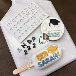 May include: A white fondant cake pop with a blue and yellow graduation cap and the text "GRAD SARAH" on a white background. A white fondant round cake topper with the text "Class of 2025" and a black graduation cap. A white silicone mold with the alphabet and numbers. The text "A HAP 222" is on the white background.