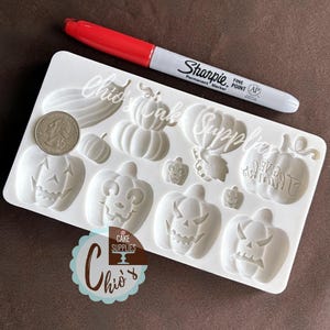 May include: A white silicone mold featuring various pumpkin designs, including jack-o'-lantern faces and pumpkin shapes. A red and silver Sharpie marker and a US quarter are next to the mold. The mold is from Chio's Cake Supplies.