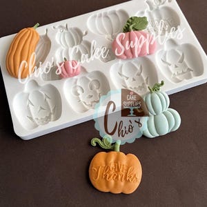 May include: A white silicone mold with pumpkin shapes and several finished fondant pumpkins in orange, pink, and blue. One orange pumpkin has the words "Give Thanks" on it. The text "Chio's Cake Supplies" is also visible.