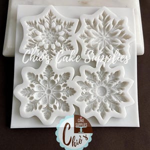 May include: Four white snowflake-shaped silicone molds for cake decorating. Each mold features a detailed, symmetrical design. The molds are on a white surface, with the text "Chio's Cake Supplies" visible. A logo with the name "Chio's" is also present.