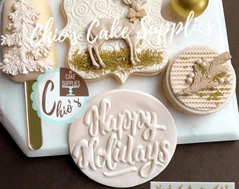 May include: A collection of decorated cookies featuring a tree-shaped cake pop, a rectangular cookie with a reindeer, a round cookie with a pine branch, and a round cookie with the text Happy Holidays, alongside a silicone mold with holiday motifs.