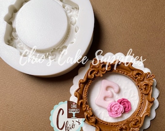 May include: A white silicone mold for making a cake topper in the shape of a decorative frame with a letter E and two pink roses. The mold is shown next to a finished cake topper made with brown fondant.