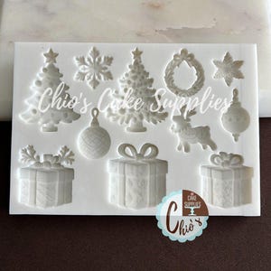 May include: A white silicone mold featuring Christmas-themed shapes. The mold includes Christmas trees, snowflakes, ornaments, gift boxes, a reindeer, and a wreath. The text "Chio's Cake Supplies" is visible on the mold.