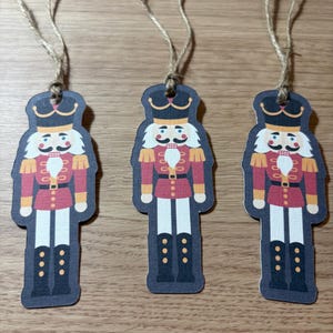 May include: Three decorative nutcracker ornaments with string hangers. Each ornament features a red and gold uniform, white hair and mustache, and black boots. The ornaments are on a light brown surface.