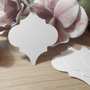 May include: White, textured paper tags in a Moroccan tile shape, with a small hole at the top for stringing. The tags are displayed on a wooden surface, with a large pink flower and green leaves in the background.