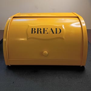 May include: A bright yellow metal bread box with a roll-top lid. The front of the container features a raised rectangular panel with the word BREAD printed in bold black capital letters and a small circular knob for opening the lid.