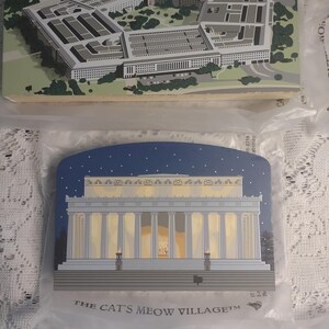 May include: Two wooden plaques featuring architectural illustrations. The top plaque depicts the Pentagon in shades of gray and green. The bottom plaque shows the Lincoln Memorial under a starry night sky. The text "THE CAT'S MEOW VILLAGE" is printed on the bottom plaque.