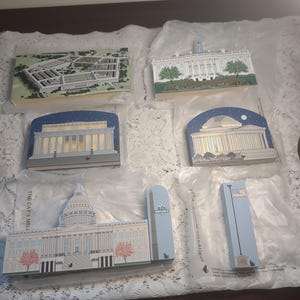 May include: A collection of rectangular wooden ornaments depicting iconic American landmarks. The ornaments include the Pentagon, White House, Capitol Building, and other monuments. Each ornament is painted with detailed architectural features and packaged in clear plastic.