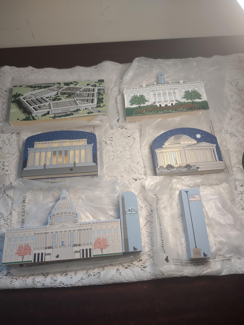 May include: A collection of rectangular wooden ornaments depicting iconic American landmarks. The ornaments include the Pentagon, White House, Capitol Building, and other monuments. Each ornament is painted with detailed architectural features and packaged in clear plastic.