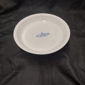 May include: White, round serving dish with a blue floral design in the center. The dish has a slightly raised edge and is set against a dark background. The floral pattern consists of three small flower clusters.