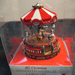 May include: A Mr. Christmas Animated Musical Carousel inside a clear plastic display box. The carousel features a red and white striped roof, a brown base with green and red railings, and several painted horses. The box base reads Mr. Christmas ANIMATED MUSICAL CAROUSEL and Try Me!