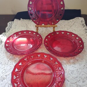 May include: A set of four red glass plates. The plates have a decorative, openwork border with triangular cutouts. One plate is displayed on a small wooden stand. The plates are arranged on a white lace tablecloth.