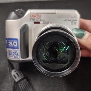 May include: A silver Olympus Camedia C-720 Ultra Zoom digital camera with an 8x optical zoom lens. The camera has a black lens cap and a black strap.