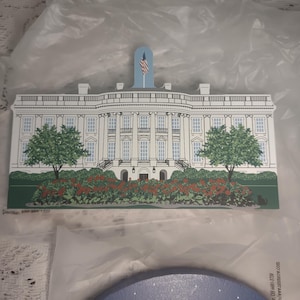May include: A wooden White House cutout with a blue flag and an American flag. The building is white with black windows, green trees, and red flowers. The cutout is on a white surface.